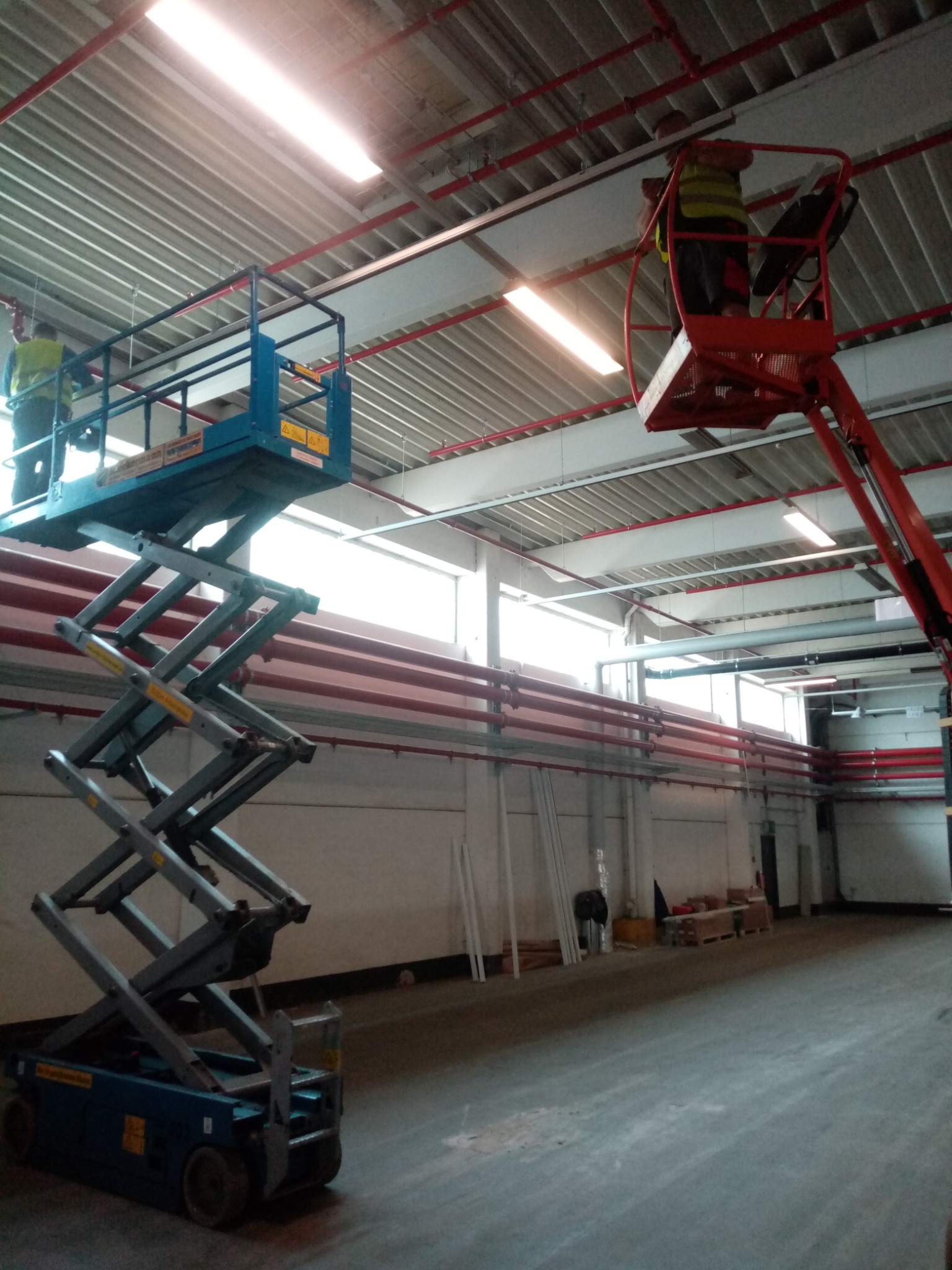 Dwóch elektryków na podnośnikach koszowych, jeden niebieski nożycowy, drugi czerwony teleskopowy, instaluje oświetlenie w hali przemysłowej z widocznymi rurami instalacyjnymi pod sufitem.