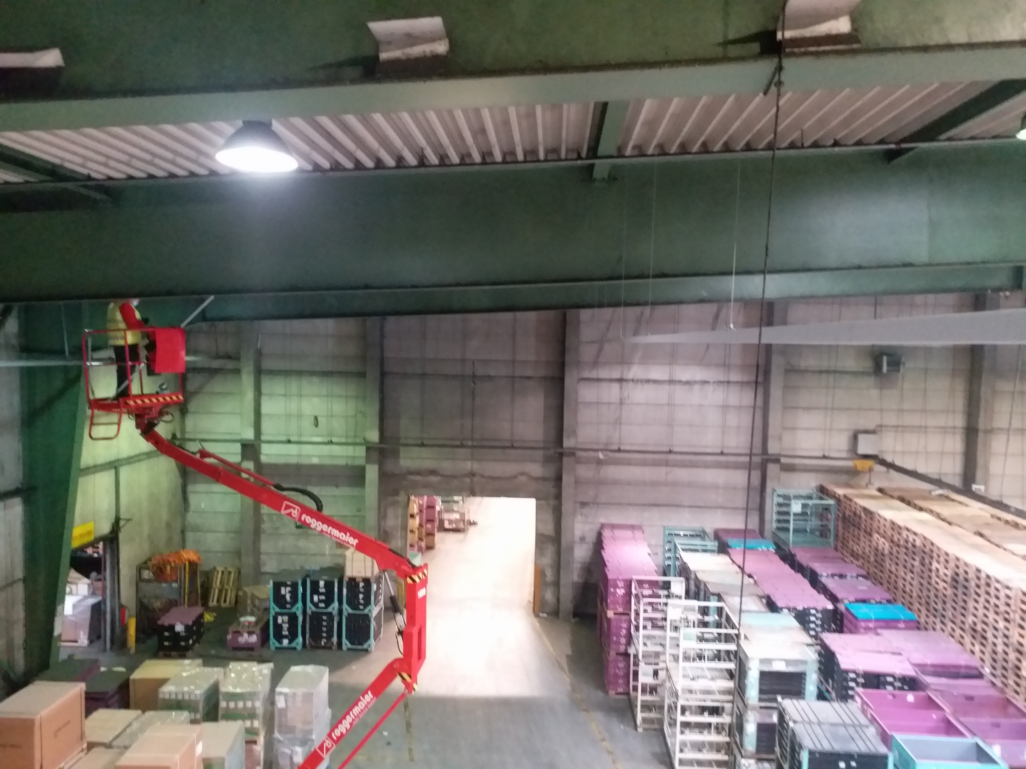 Pracownik na podnośniku koszowym, w hali magazynowej, prawdopodobnie podczas wymiany lub naprawy oświetlenia sufitowego.