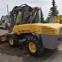 Żółto-szara koparko-ładowarka Mecalac 45X z łyżką z przodu, zaparkowana na placu budowy z widocznymi innymi pojazdami w tle.