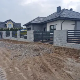 Nowoczesne ogrodzenie z szarego betonu i metalowych przęseł w kolorze antracytowym, z bramą wjazdową, na tle nowo wybudowanych domów i ziemi z gruzem.