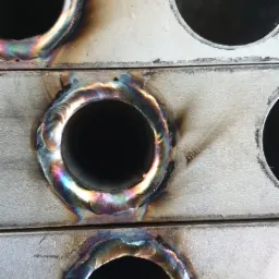 Metalowe płyty z okrągłymi otworami, widoczne ślady spawania z opalizującymi przebarwieniami termicznymi wokół otworów.