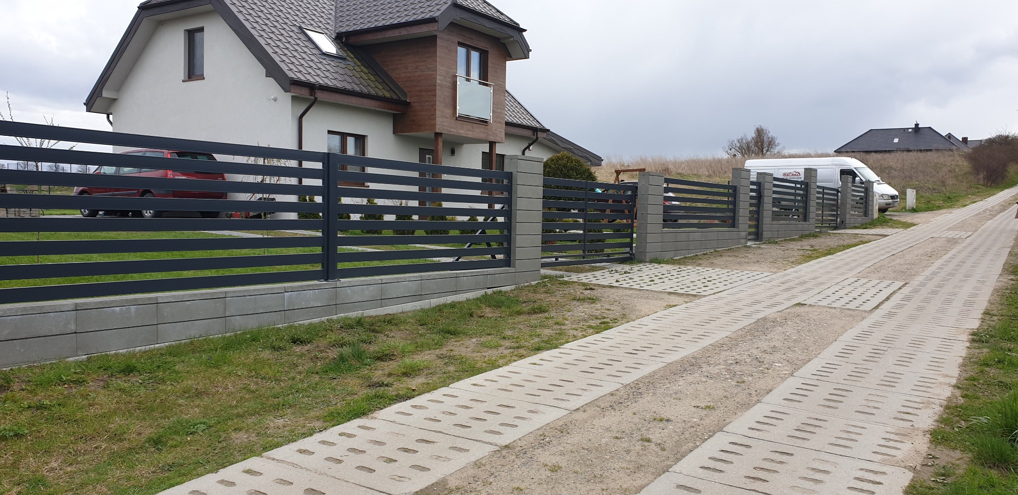 Nowoczesne ogrodzenie posesji z poziomych, grafitowych profili stalowych na betonowej podmurówce i słupkach, widok z perspektywy wjazdu na posesję, w tle dom jednorodzinny i samochód dostawczy.