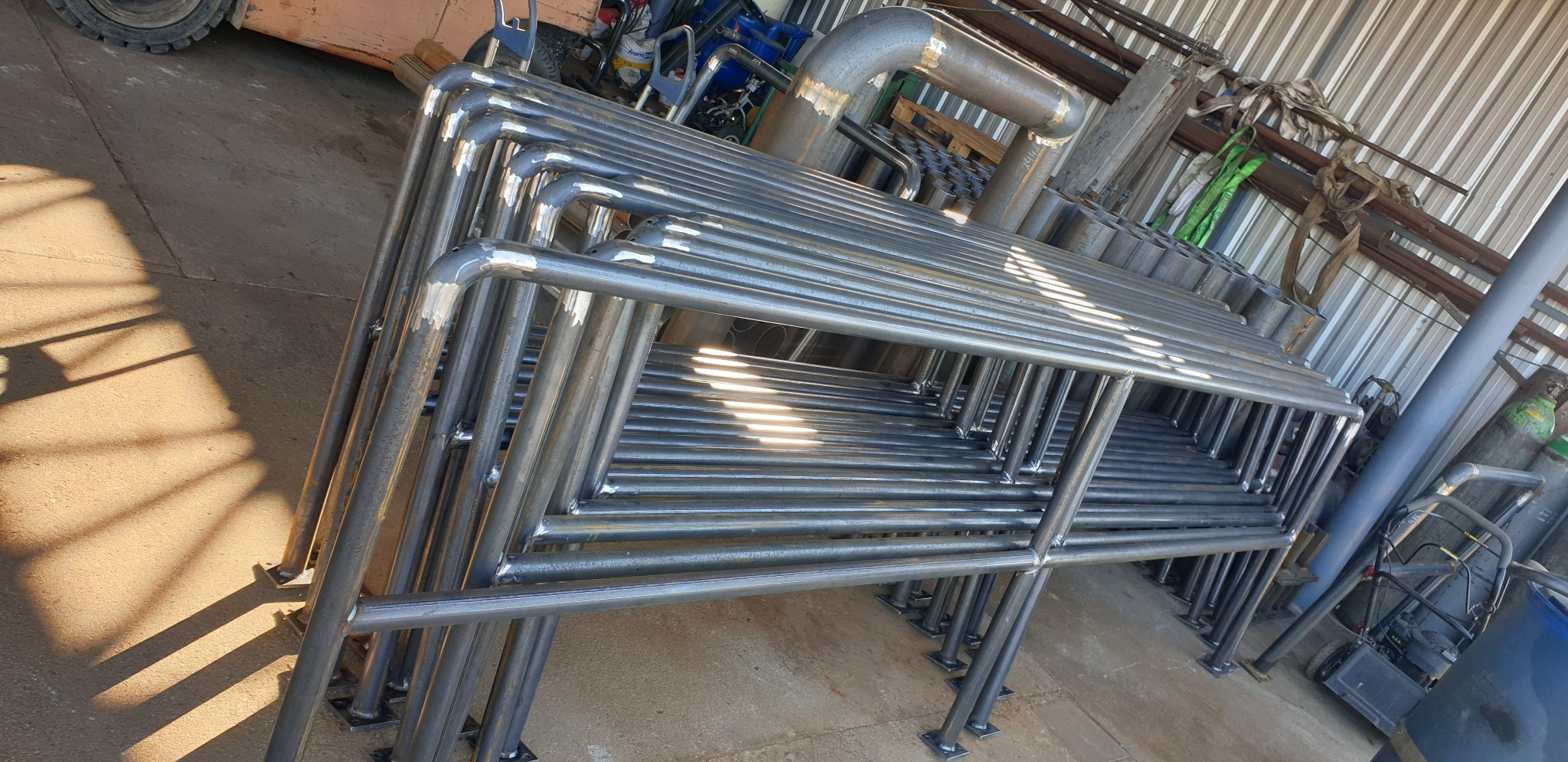 Stos metalowych barierek, świeżo po spawaniu, z widocznymi śladami obróbki, ułożonych w warsztacie, z elementami wyposażenia w tle.