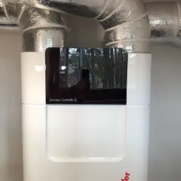 Biała centrala rekuperacyjna Zehnder ComfoAir Q z widocznymi izolowanymi przewodami wentylacyjnymi i odbiciem osoby robiącej zdjęcie w czarnym panelu.