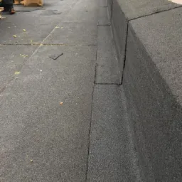 Widok z góry na płaski dach pokryty czarną papą, z widocznym fragmentem obróbki blacharskiej przy krawędzi dachu. W tle widoczne palety i butla gazowa.