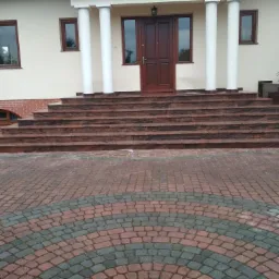 Wejście do domu z szerokimi schodami z czerwonego granitu, kolumny podtrzymujące zadaszenie, bruk na podjeździe ułożony w p&oacute;łkoliste wzory z czerwonej i szarej kostki.