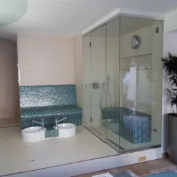 Nowoczesna łazienka z przeszklonym prysznicem, mozaikową ławką i dwiema ceramicznymi umywalkami na podłodze, uzupełniona rośliną doniczkową w szarej osłonce.