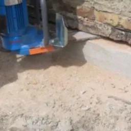 Niebieska maszyna do osuszania murów w trakcie pracy, widoczna ściana z cegieł i piasek.