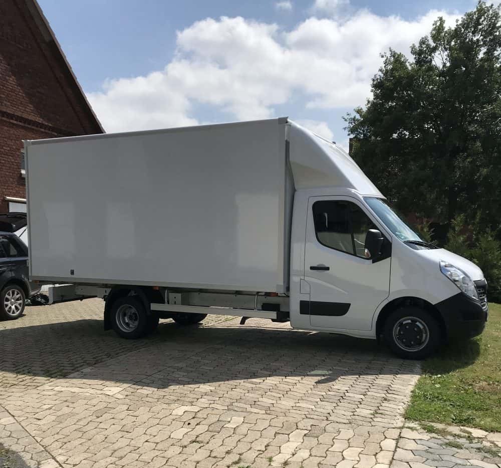 Biały samochód dostawczy typu furgon z wysoką kabiną, zaparkowany na brukowanym podjeździe przed budynkiem z czerwonej cegły w słoneczny dzień.