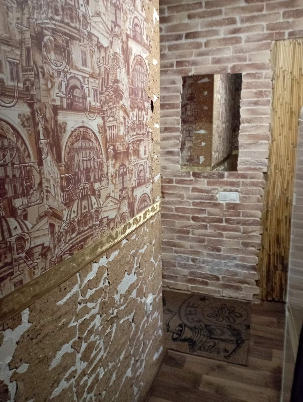 Korytarz z dekoracyjną cegłą, tapetą z motywem architektonicznym i fragmentami odsłoniętej struktury ściany, widoczny dywanik z nadrukiem.