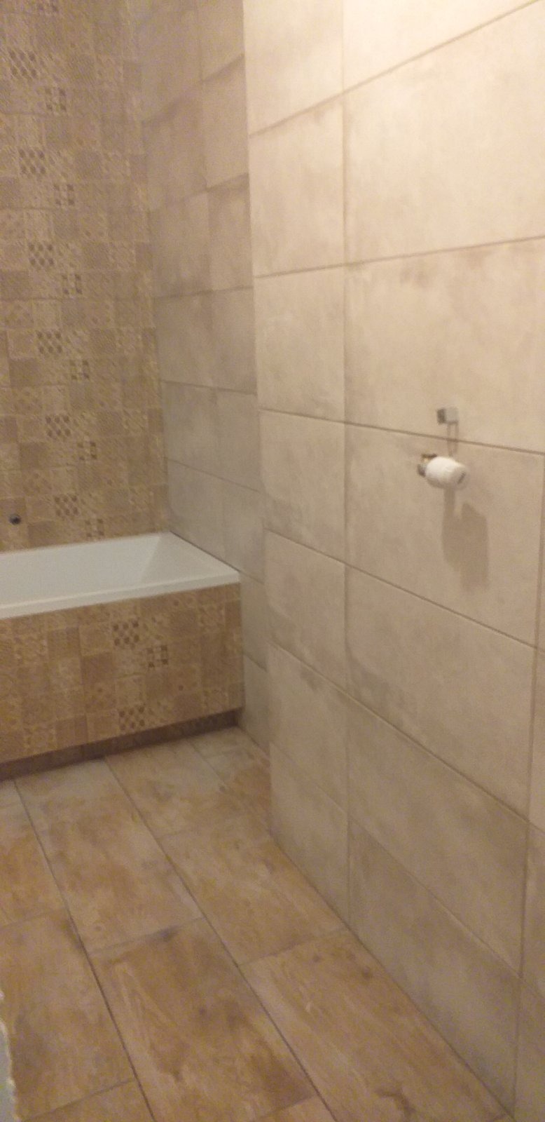 Wykończona łazienka z beżowymi płytkami na ścianach i podłodze, widoczna wanna z obudową z płytek dekoracyjnych, uchwyt na papier toaletowy.