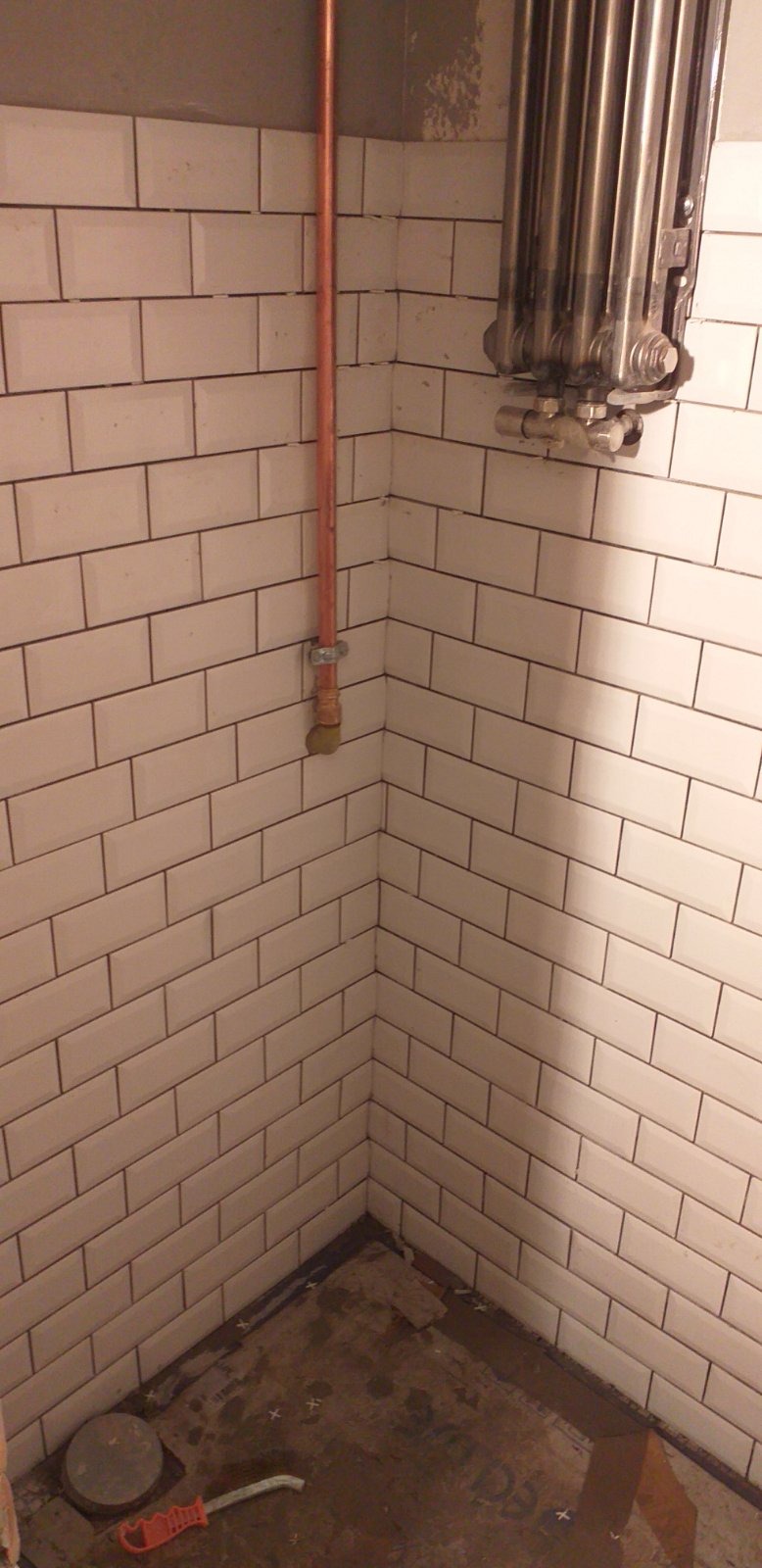 Wnętrze łazienki w Warszawie w trakcie remontu: białe, prostokątne płytki ułożone na ścianach, miedziana rurka instalacji, fragment grzejnika i narzędzia na podłodze.