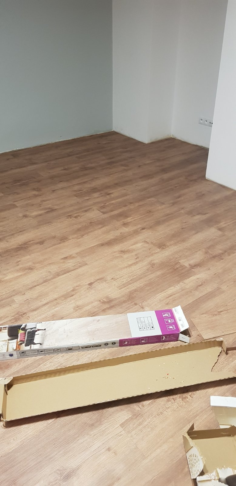 Układanie paneli podłogowych w pokoju, widoczna częściowo ułożona podłoga, karton z panelami oraz puste opakowanie po panelach.