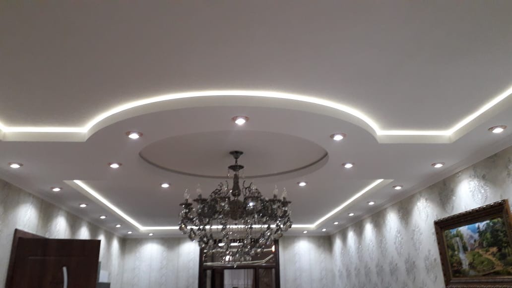 Sufit podwieszany z oświetleniem LED w kształcie okręgów i prostokątów, centralnie zawieszony ozdobny żyrandol, tapeta w delikatny wzór, obraz na ścianie.