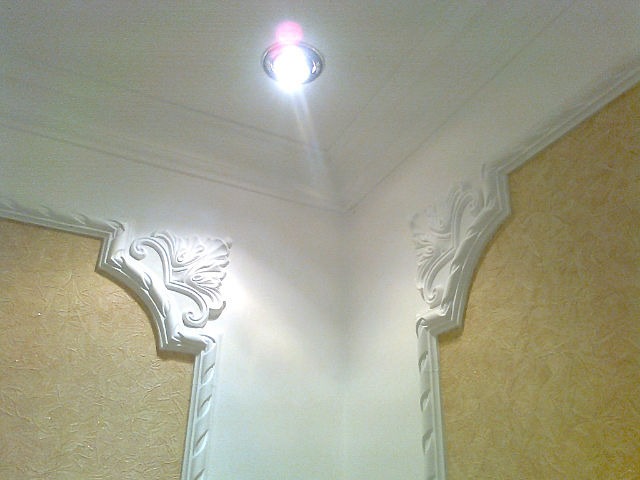 Dekoracyjny narożnik sufitu z gipsowymi ornamentami w stylu retro, kremowa ściana z teksturą i punktowe oświetlenie sufitowe.
