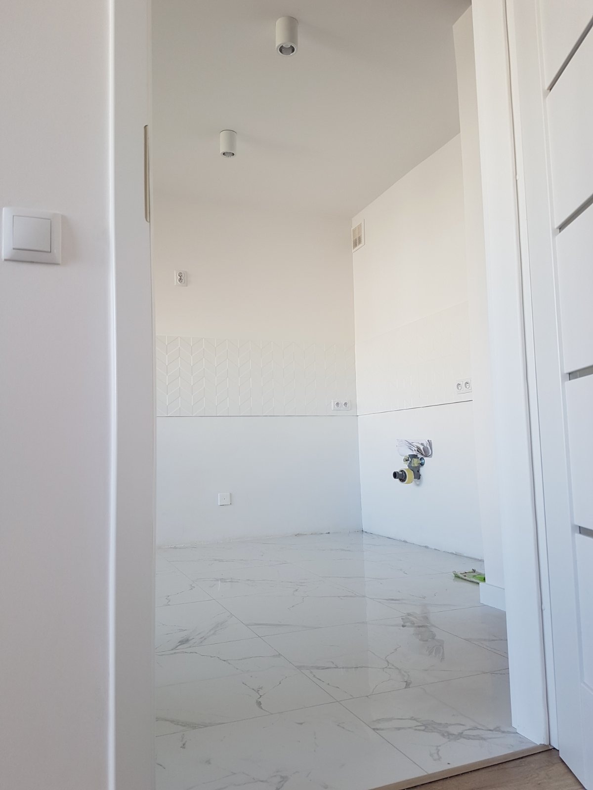 Wnętrze łazienki w trakcie remontu, widoczne białe płytki na podłodze imitujące marmur, częściowo wykończone ściany z dekoracyjną białą mozaiką, przygotowane przyłącza wodno-kanalizacyjne, surowe...