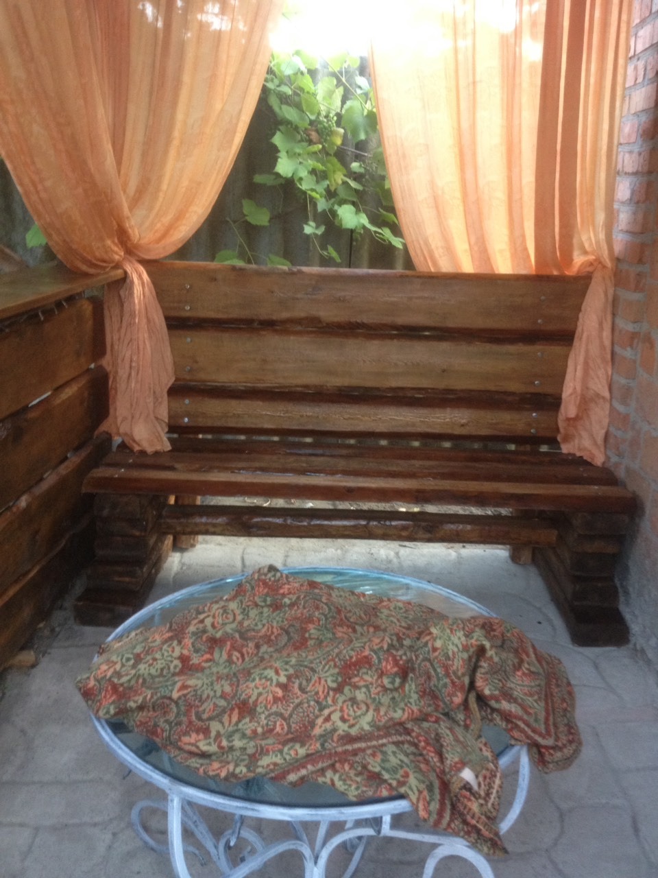 Zadaszone patio z drewnianą ławką, zwisającymi pomarańczowymi zasłonami i stolikiem z wzorzystym pledem, widok na zieleń za ławką.