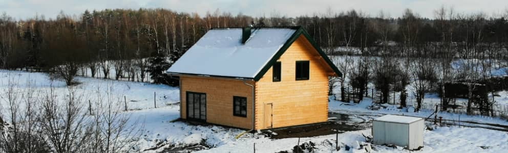 Drewniany dom z zielonym dachem pokryty śniegiem, otoczony zimowym krajobrazem z lasem w tle.