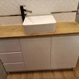 Łazienka z białą, prostokątną umywalką nablatową i czarną baterią, umieszczona na szafce z białymi, lakierowanymi frontami i blatem o strukturze jasnego drewna.