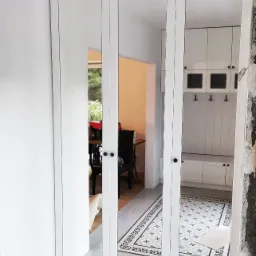 Biała szafa wnękowa z lustrami, odbijająca fragment przedpokoju z wzorzystą podłogą i widokiem na jadalnię przez otwarte drzwi.