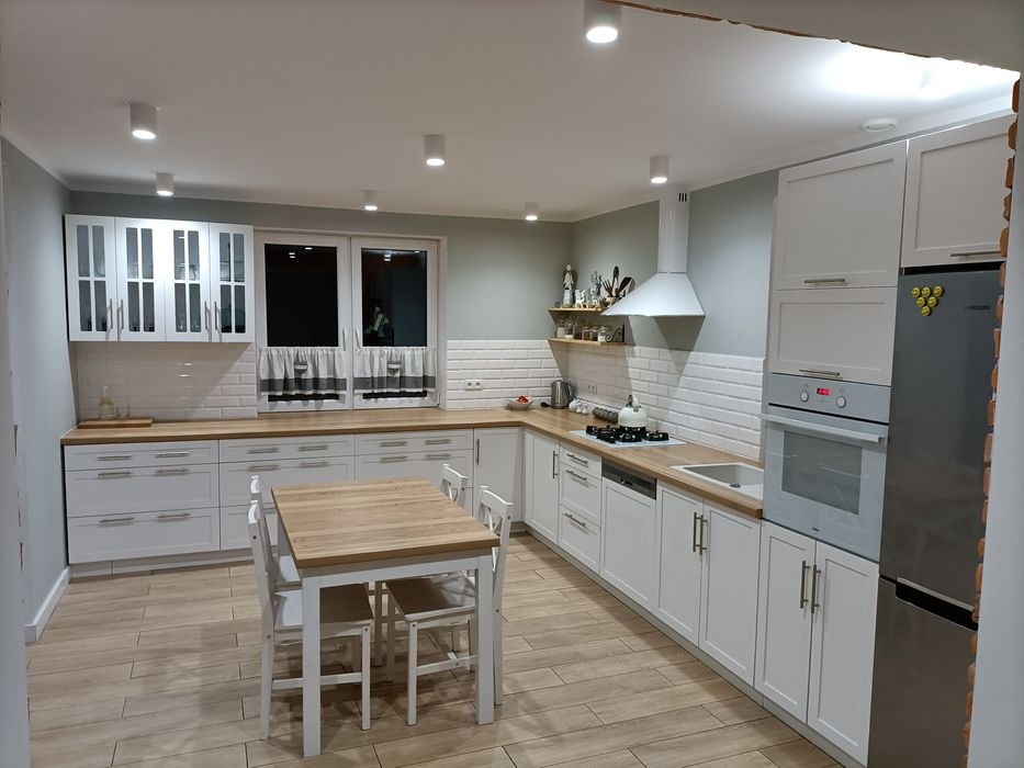 Jasna kuchnia z białymi frontami, drewnianym blatem i stołem z krzesłami, oświetlona reflektorami sufitowymi, z okapem i płytkami imitującymi cegłę na ścianie.