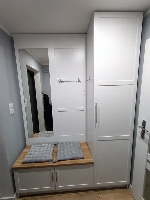 Biała zabudowa przedpokoju z siedziskiem z drewnianym blatem, lustrem i wieszakami, uzupełniona szarymi poduszkami.