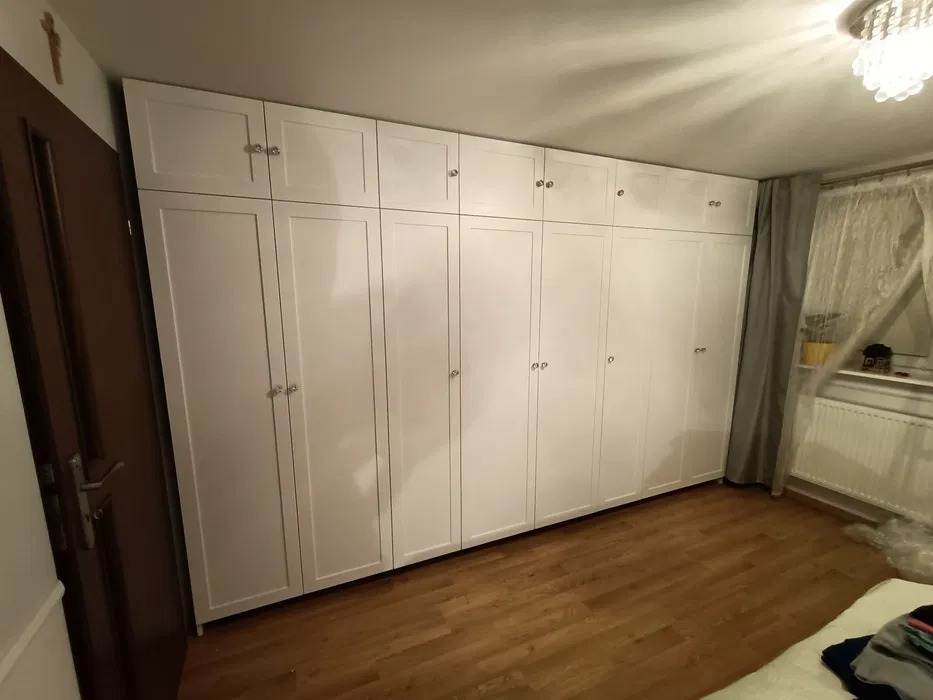 Biała szafa na wymiar z górnymi szafkami, zamontowana od podłogi do sufitu w Ciechanowie, widoczna z perspektywy dolnej, z drewnianą podłogą i fragmentem okna z zasłoną.