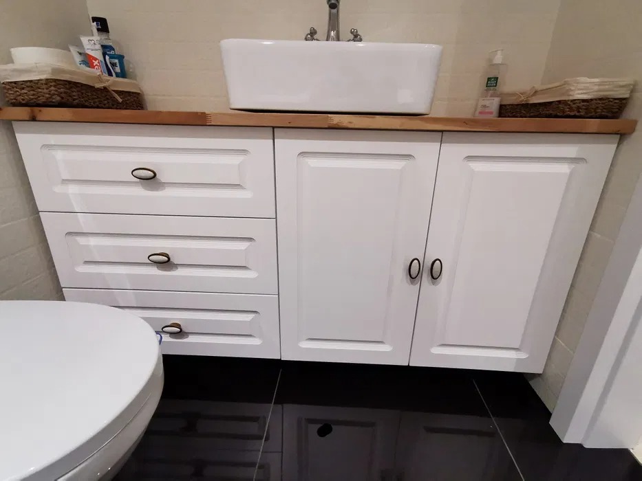 Biała szafka łazienkowa z trzema szufladami i dwojgiem drzwiczek, drewniany blat z nablatową umywalką, czarna podłoga z połyskiem, widoczny fragment toalety.