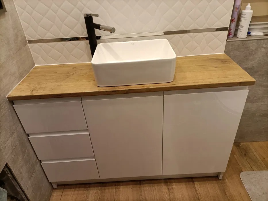 Łazienka z białą, prostokątną umywalką nablatową i czarną baterią, umieszczona na szafce z białymi, lakierowanymi frontami i blatem o strukturze jasnego drewna.