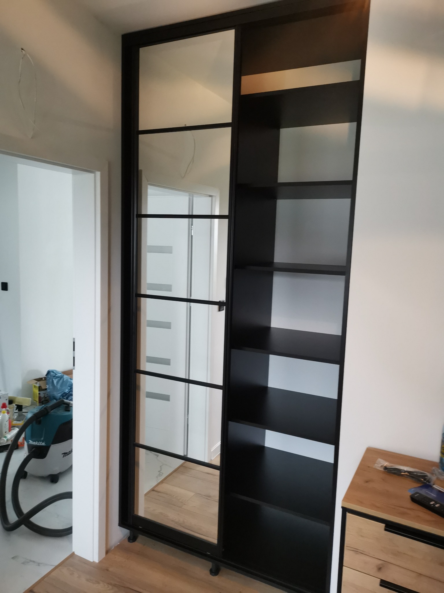 Czarna szafa z lustrzanymi drzwiami przesuwnymi, widoczne półki, w tle odkurzacz i fragment komody.