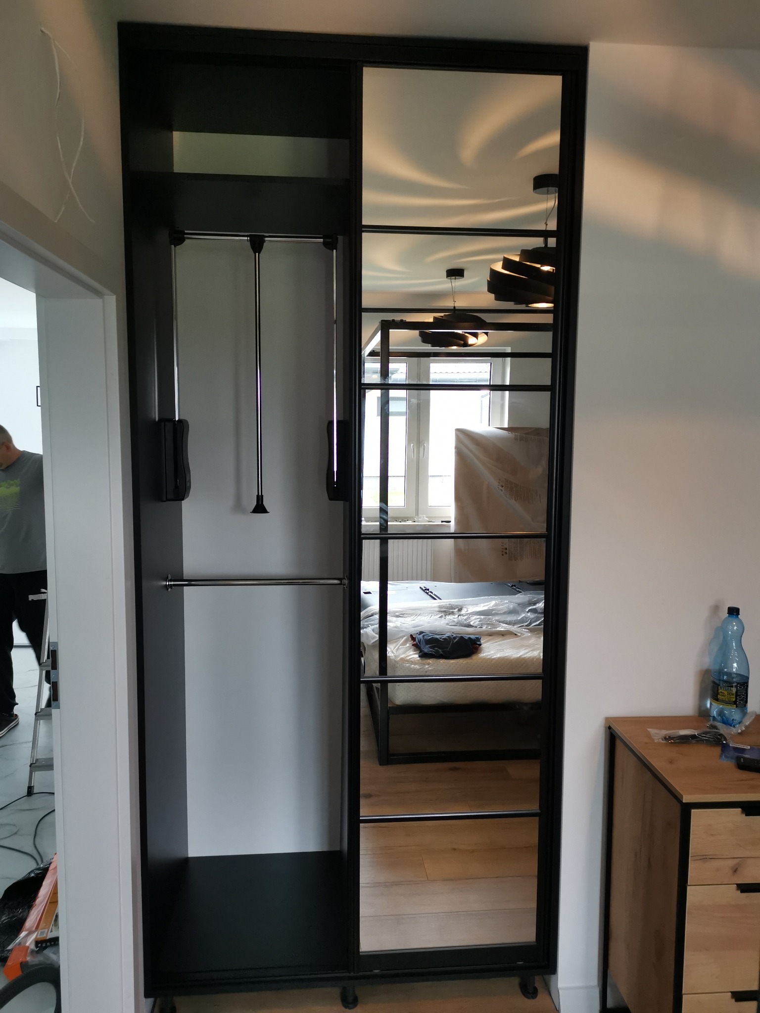 Czarna szafa przesuwna z lustrem, widoczne wnętrze z drążkami i windą na ubrania, odbicie pokoju z łóżkiem i lampami.