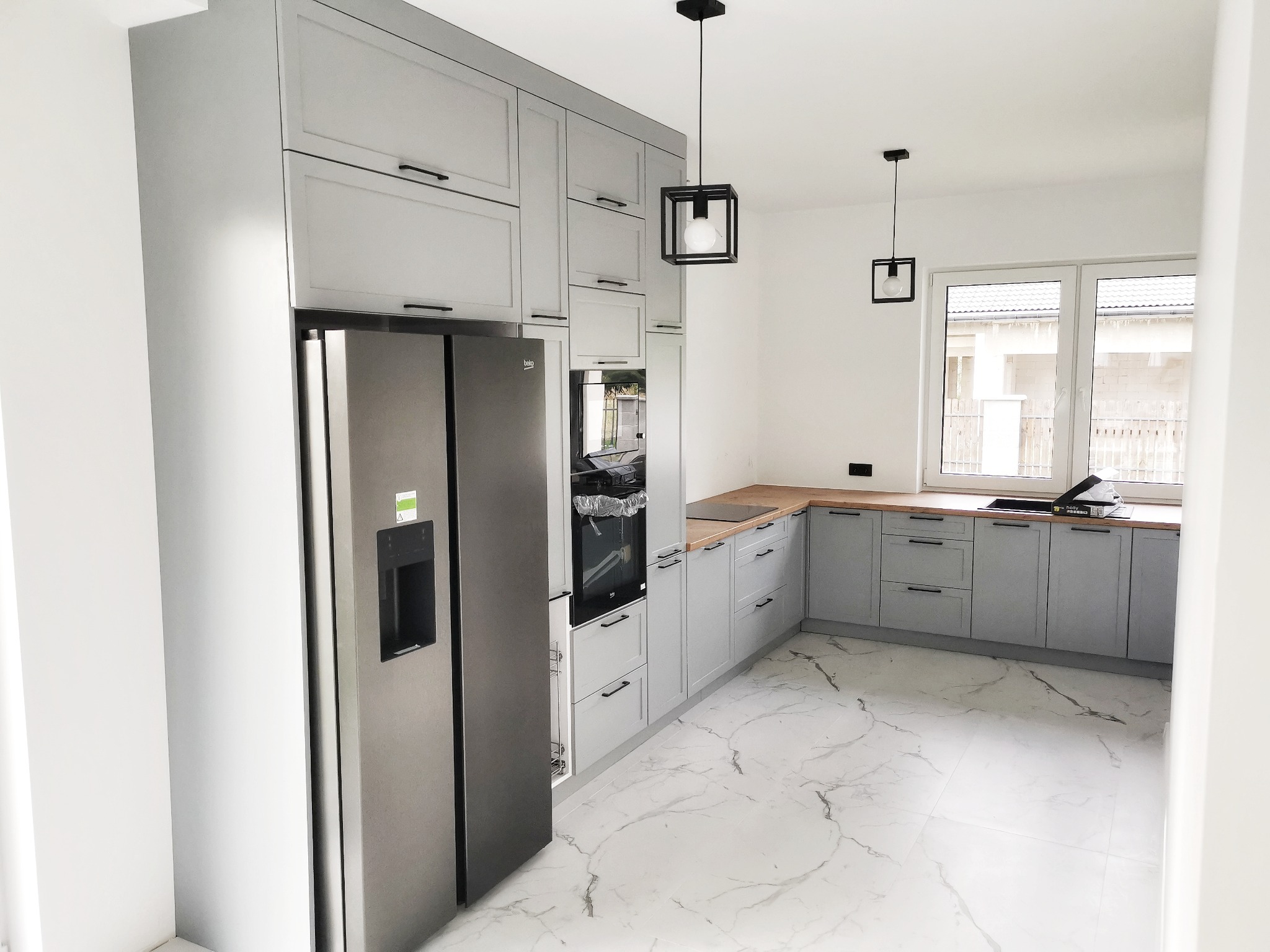 Jasnoszara zabudowa kuchenna z drewnianym blatem, nowoczesne lampy wiszące, marmurowe płytki na podłodze, widok na okno.