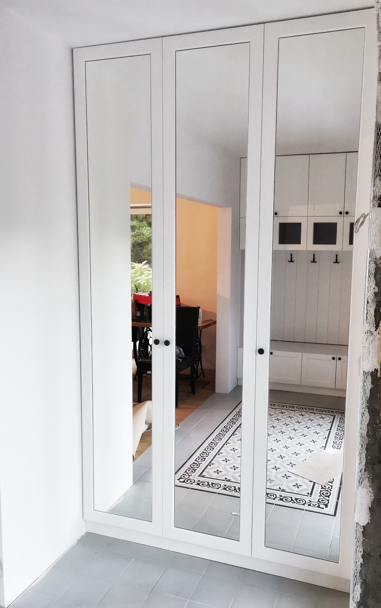 Biała szafa wnękowa z lustrami, odbijająca fragment przedpokoju z wzorzystą podłogą i widokiem na jadalnię przez otwarte drzwi.