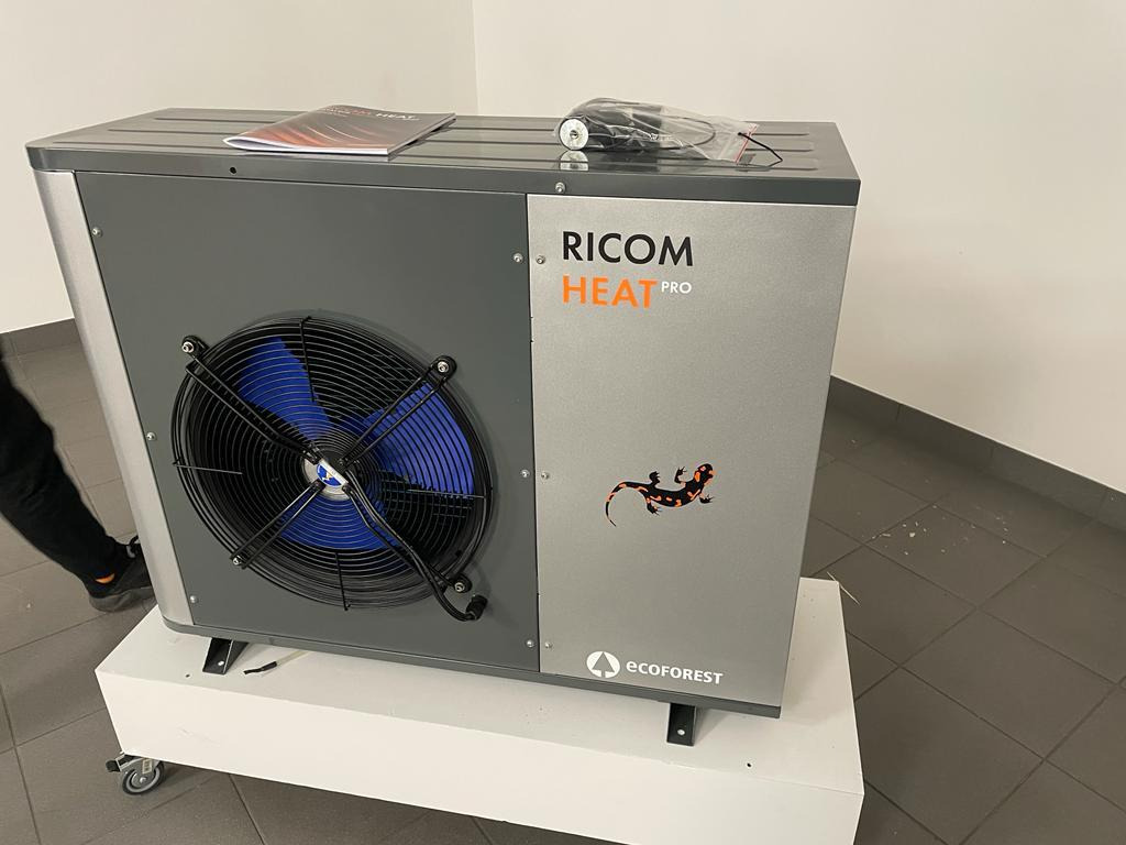 Szaro-srebrna pompa ciepła Ricom Heat Pro na białym podwyższeniu z kółkami, widoczny wentylator z niebieskimi łopatkami i naklejka z sylwetką jaszczurki.