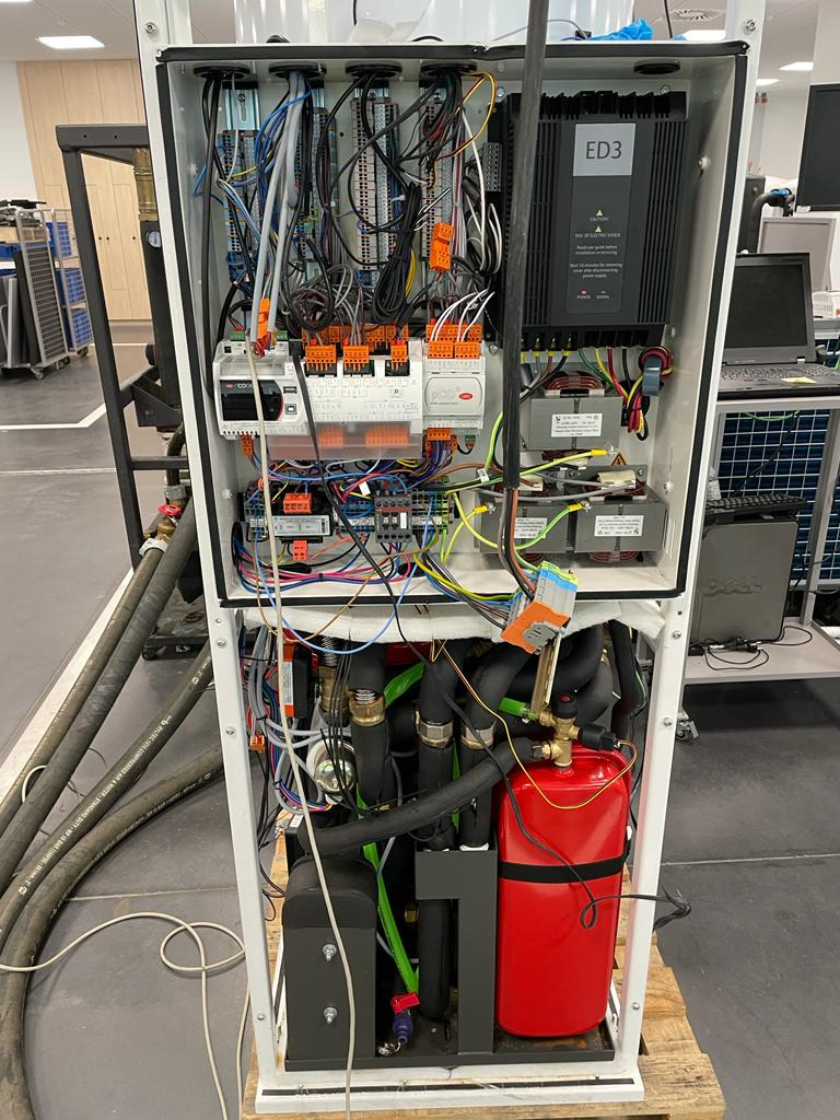 Otwarta obudowa pompy ciepła z widocznymi elementami sterowania, przewodami i instalacją hydrauliczną, w tle widoczny sprzęt komputerowy i wózki transportowe.