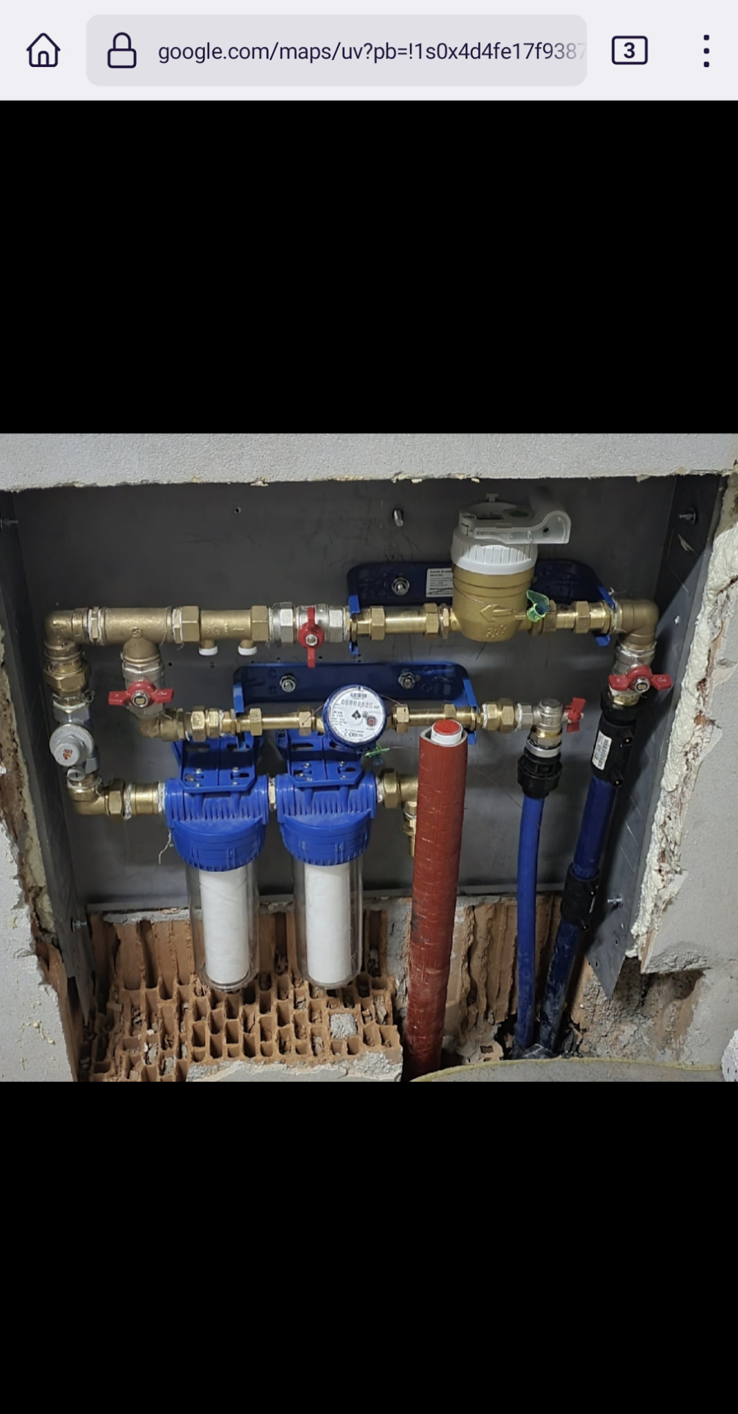 Zamontowany system filtracji wody z dwoma cylindrycznymi filtrami, manometrem, licznikiem wody i rurami w ścianie z widoczną cegłą.