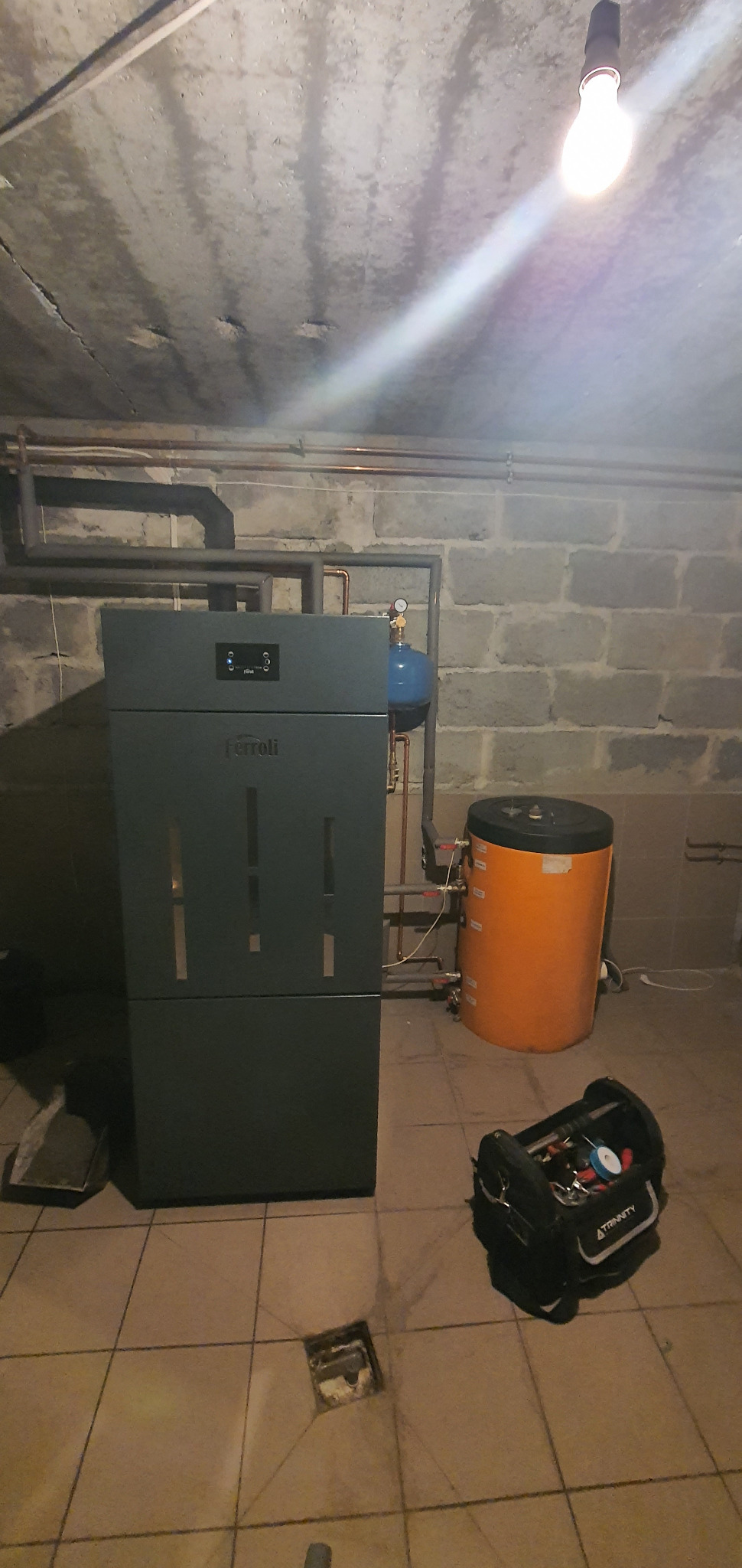 Szary piec Ferroli z podłączonym niebieskim zbiornikiem wyrównawczym i pomarańczowym zasobnikiem ciepłej wody, widoczne rury instalacji oraz skrzynka z narzędziami na podłodze z płytek, surowy...