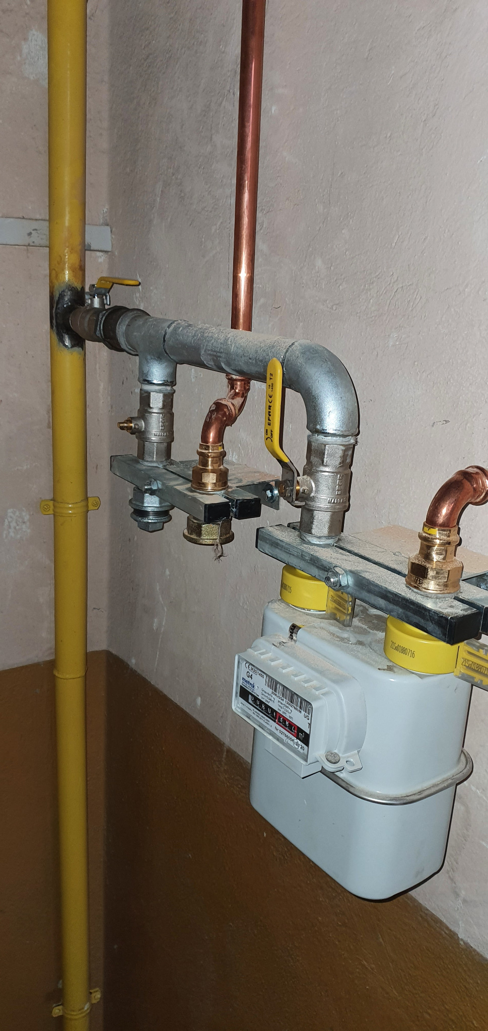 Instalacja gazowa z licznikiem, rurami miedzianymi i zaworami kulowymi, zamontowana przy ścianie z widocznymi śladami użytkowania.