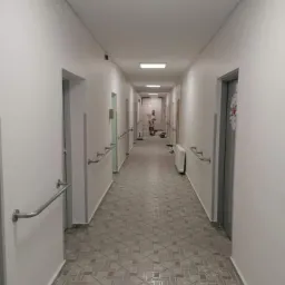 Długi korytarz w budynku, pomalowany na biało, z szarymi drzwiami i poręczami. Na końcu korytarza widoczna osoba malująca ścianę przy użyciu drabiny.