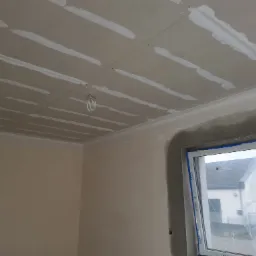 Płyty karton-gips na suficie, widoczne łączenia zaszpachlowane, przygotowane do dalszych prac wykończeniowych, okno zabezpieczone folią.