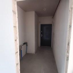 ReMalFach - Surowy stan mieszkania po wylewce, widoczne instalacje wodne, ościeżnica w trakcie obróbki, czarne drzwi wejściowe w głębi.
