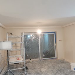 ReMalFach - Wnętrze w trakcie remontu w Gnieźnie: widoczny sufit z dekoracyjną listwą, ściany przygotowane do malowania, okna zabezpieczone folią, oświetlenie robocze i drabina malarska.