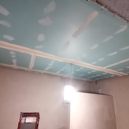 ReMalFach - Montaż płyt gipsowo-kartonowych na suficie, widoczne łączenia wypełnione masą szpachlową przed szlifowaniem, surowy stan wykończenia wnętrza z widocznym oknem i instalacją sanitarną w tle.