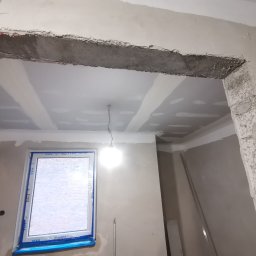 ReMalFach - Surowy otwór drzwiowy z odsłoniętym betonem i siatką wzmacniającą, widoczne płyty kartonowo-gipsowe na suficie z nałożoną masą szpachlową, okno z niebieską folią zabezpieczającą, surowe ściany...