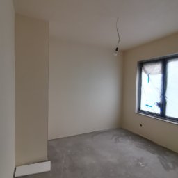 ReMalFach - Wnętrze po gładziowaniu i malowaniu na beżowo. Okno zabezpieczone folią. Podłoga szara, surowa. Wystający fragment ściany. Wisząca żarówka.