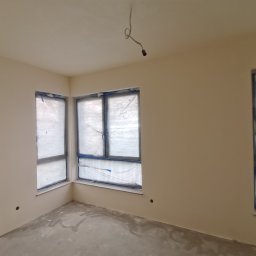 ReMalFach - Wnętrze pokoju po gładziowaniu, z oknami zabezpieczonymi folią. Ściany pomalowane na jasny kolor, widoczny surowy betonowy filar.