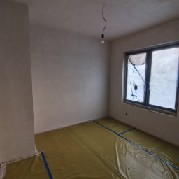 ReMalFach - Pokój po szpachlowaniu, zabezpieczony folią. Widoczne gładkie ściany, okno osłonięte folią i wisząca żarówka. Przygotowanie do dalszych prac wykończeniowych.