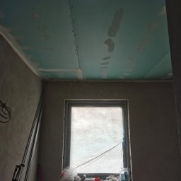ReMalFach - Płyty KG na suficie z widocznymi łączeniami przed szpachlowaniem w pomieszczeniu z oknem i surowymi ścianami. Widoczne profile i narzędzia.