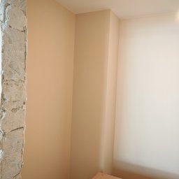 ReMalFach - Wykończony róg pokoju po pracach w Gniezno: gładkie, beżowe ściany kontrastują z surowym betonem futryny. Na podłodze kartony z materiałami wykończeniowymi.