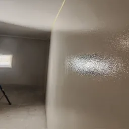 Świeżo pomalowana, błyszcząca ściana w neutralnym odcieniu, oświetlona lampą LED na statywie. Widoczne ślady po taśmie malarskiej przy suficie.